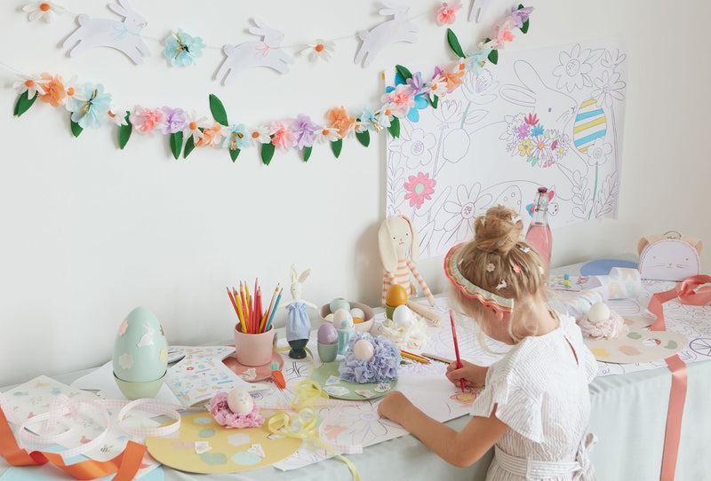 A young girl draws at a table covered with Easter eggs and stickers, with a Easter colouring poster above and floral garlands.