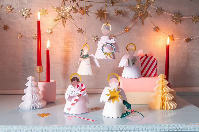 Paper angel ornaments stand on the table and some hang from the ceiling along with a star garland. Honeycomb trees are placed beside lighted red taper candles.