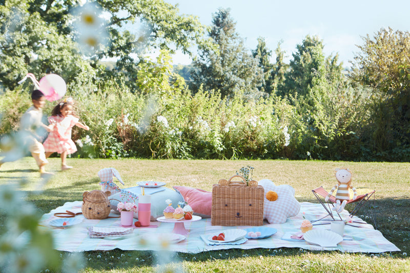 In a sunny garden, two young children run in the background whilst a picnic is laid in front of them featuring bear wicker baskets, gingham cushions and scallop-edged plates.