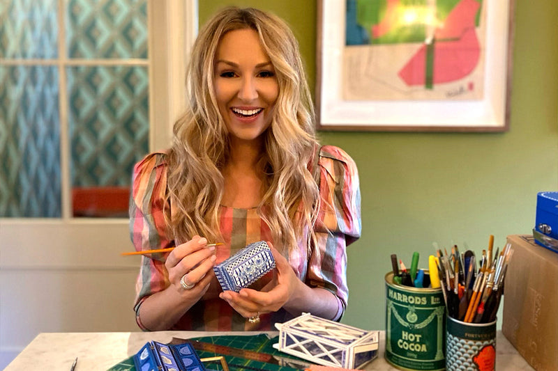 Lucy Clayton smiles at the camera whilst holding a paintbrush and trinket box, with other craft supplies on the table in front of her.