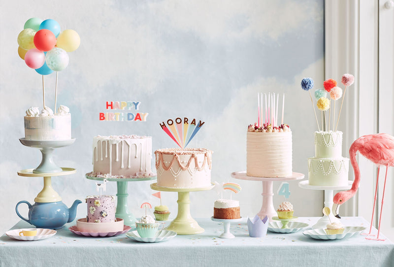 A line of large cakes covered in frosting placed on cake stands display a range of colourful candles and cake toppers.
