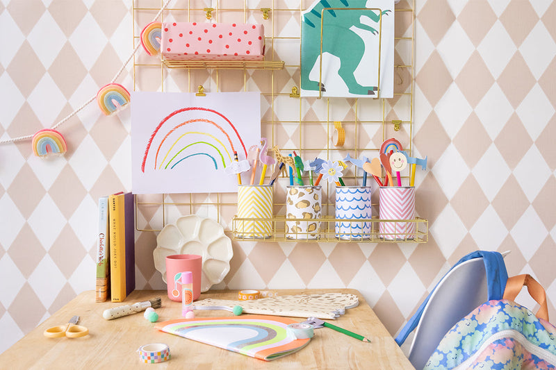 A gold rack holds 4 desk tidys which have pencils inside, and a desk has a notebook and other craft supplies.