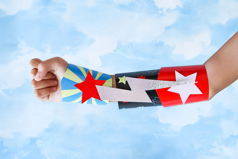 In front of a background of the sky, an arm displays a DIY superhero cuff made with coloured card and finished with stars and a lightning bolt.