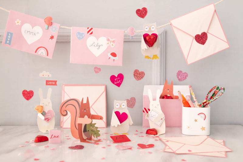 Valentine's envelopes in pink and white with hearts hang from a banner above Hugger crafts created in designs of woodland animals such as squirrels and bunnies. Glitter heart stickers and felt heart hair slides are also placed amongst the cards.