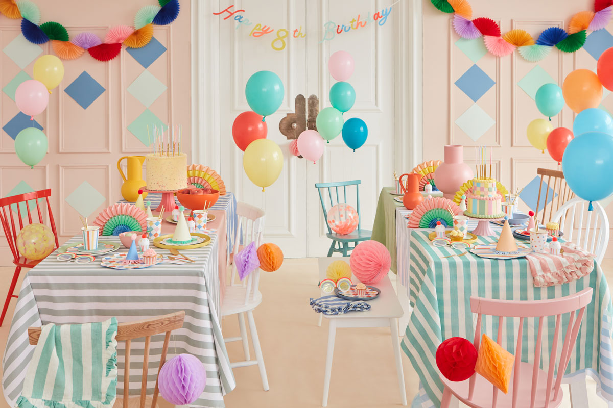 A dining room displays a multitude of colorful garlands and tableware, including honeycomb ornaments, pastel balloons, and ruffled-edge napkins.