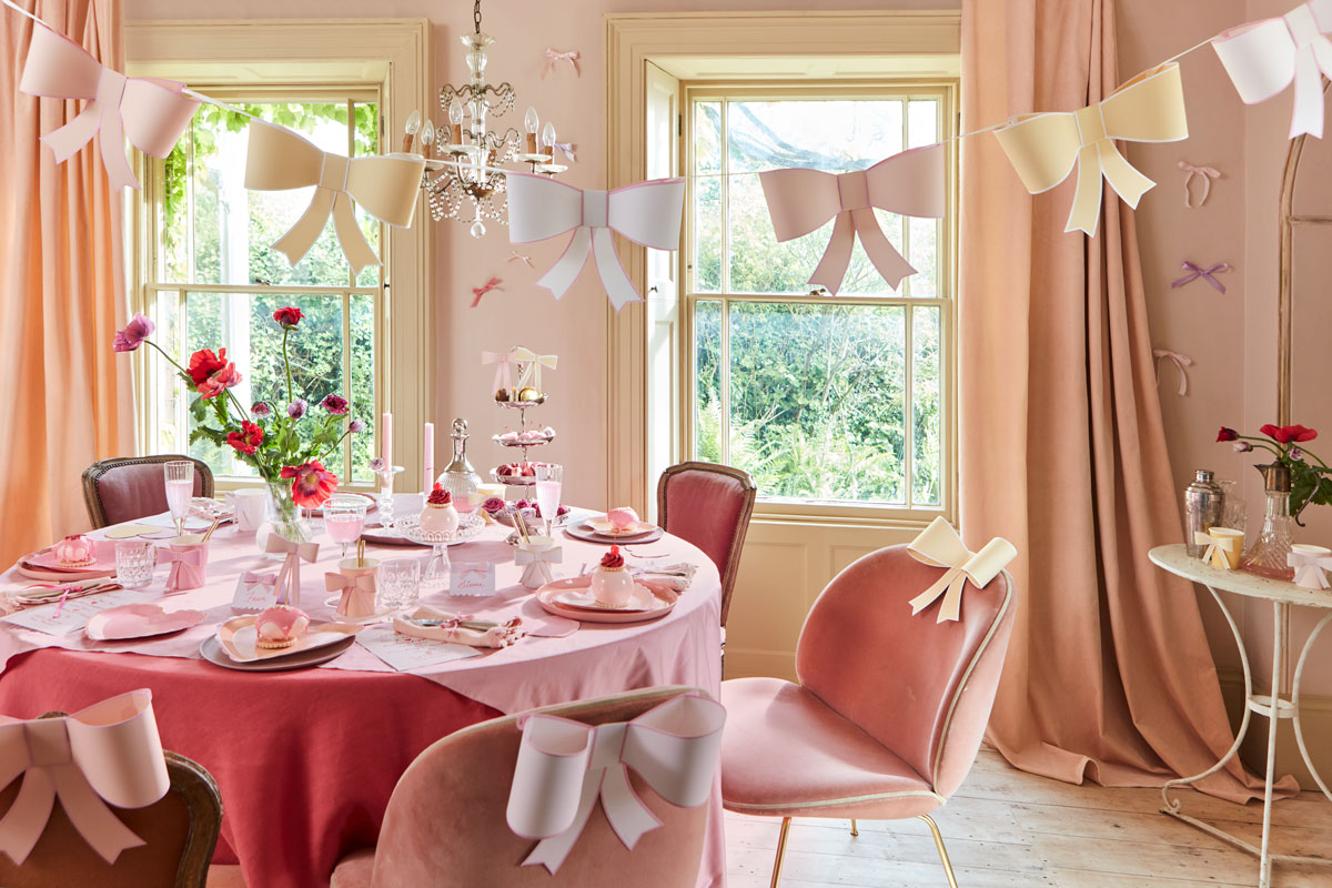 In a pink-themed dining room, a string of bows in pastel shades of pink, yellow, and blue hangs in the foreground whilst a table surrounded by dusty pink chairs is bedecked in pink heart plates and tableware accessorized with bows.